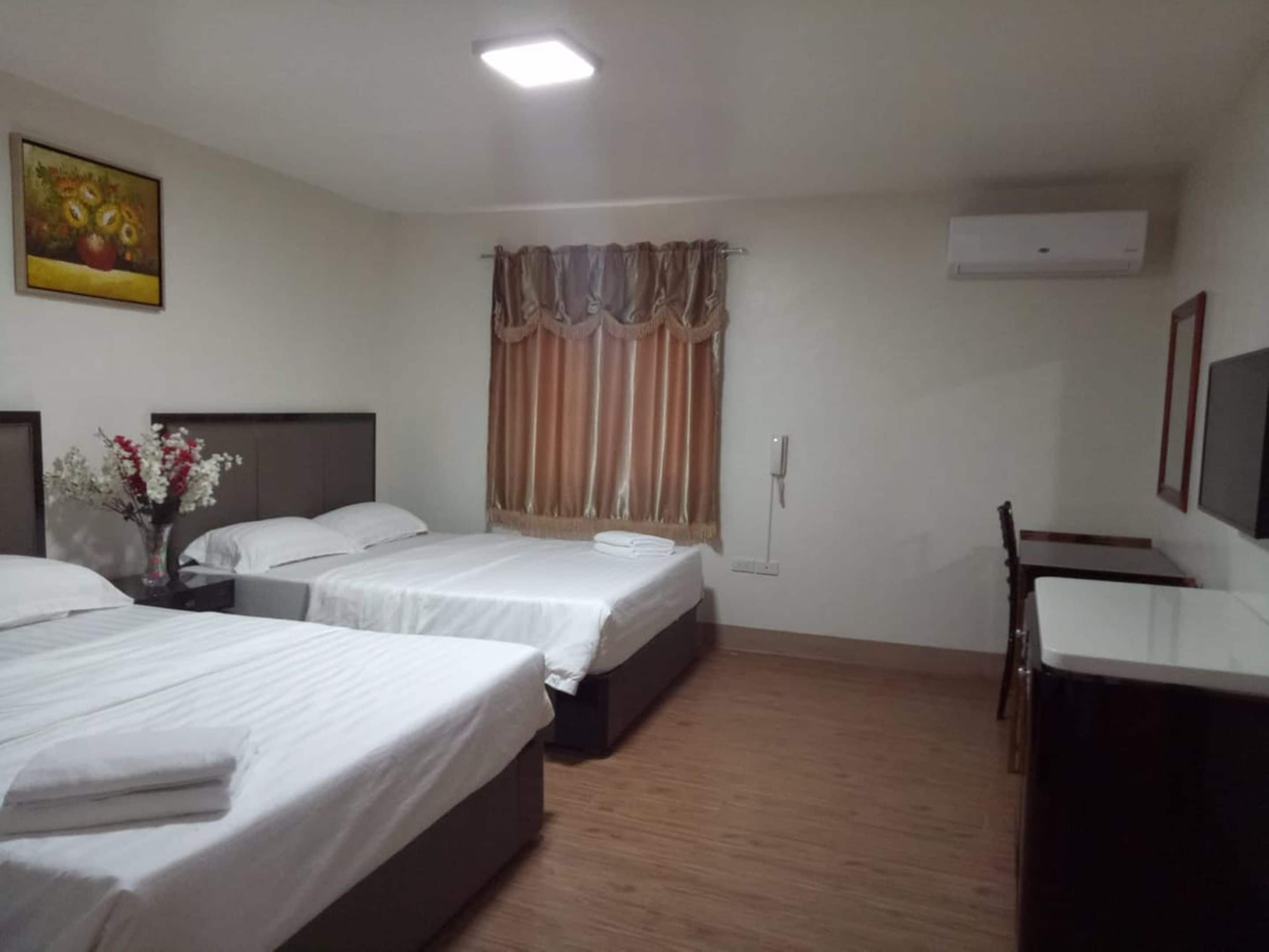 A hotel room with three single beds, a desk, a chair, a vase with flowers, a painting, a window with curtains, and an air conditioning unit.