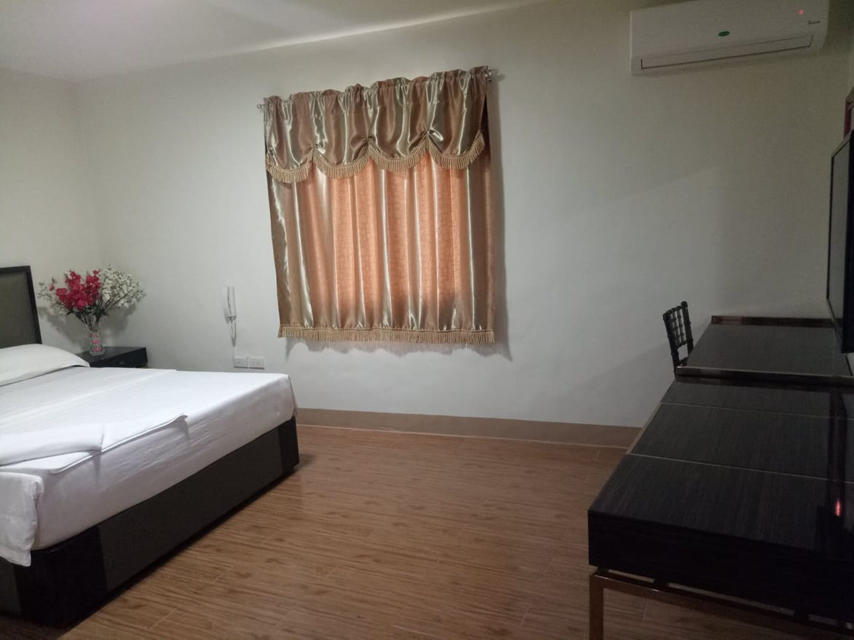 Ein Hotelzimmer mit Bett, kleinem Tisch, Stuhl, Nachttisch, einer Vase mit Blumen und einem Fenster mit Vorhängen.