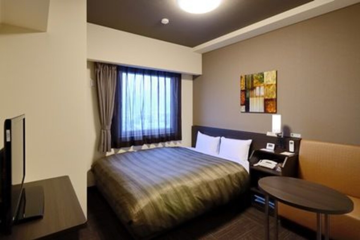 Habitación de hotel con cama, escritorio, silla, televisor y ventana con cortinas.