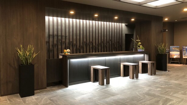 A modern reception desk with a dark wood finish, a glass partition, and a potted plant on the left. - Hotel Route Inn Grand Muroran (Muroran)