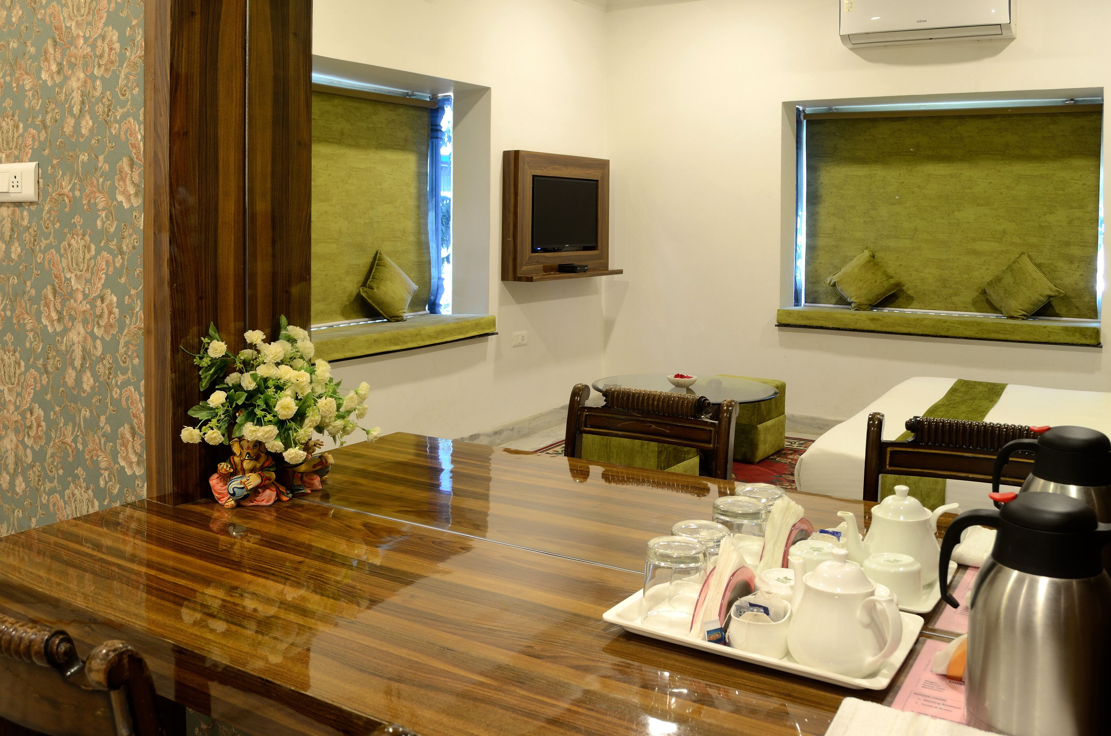 A hotel room with a wooden dining table, a flower vase, a TV mounted on the wall, and a bed with a green bedspread.