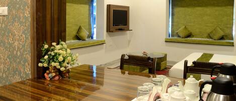 A hotel room with a wooden dining table, a flower vase, a TV mounted on the wall, and a bed with a green bedspread.