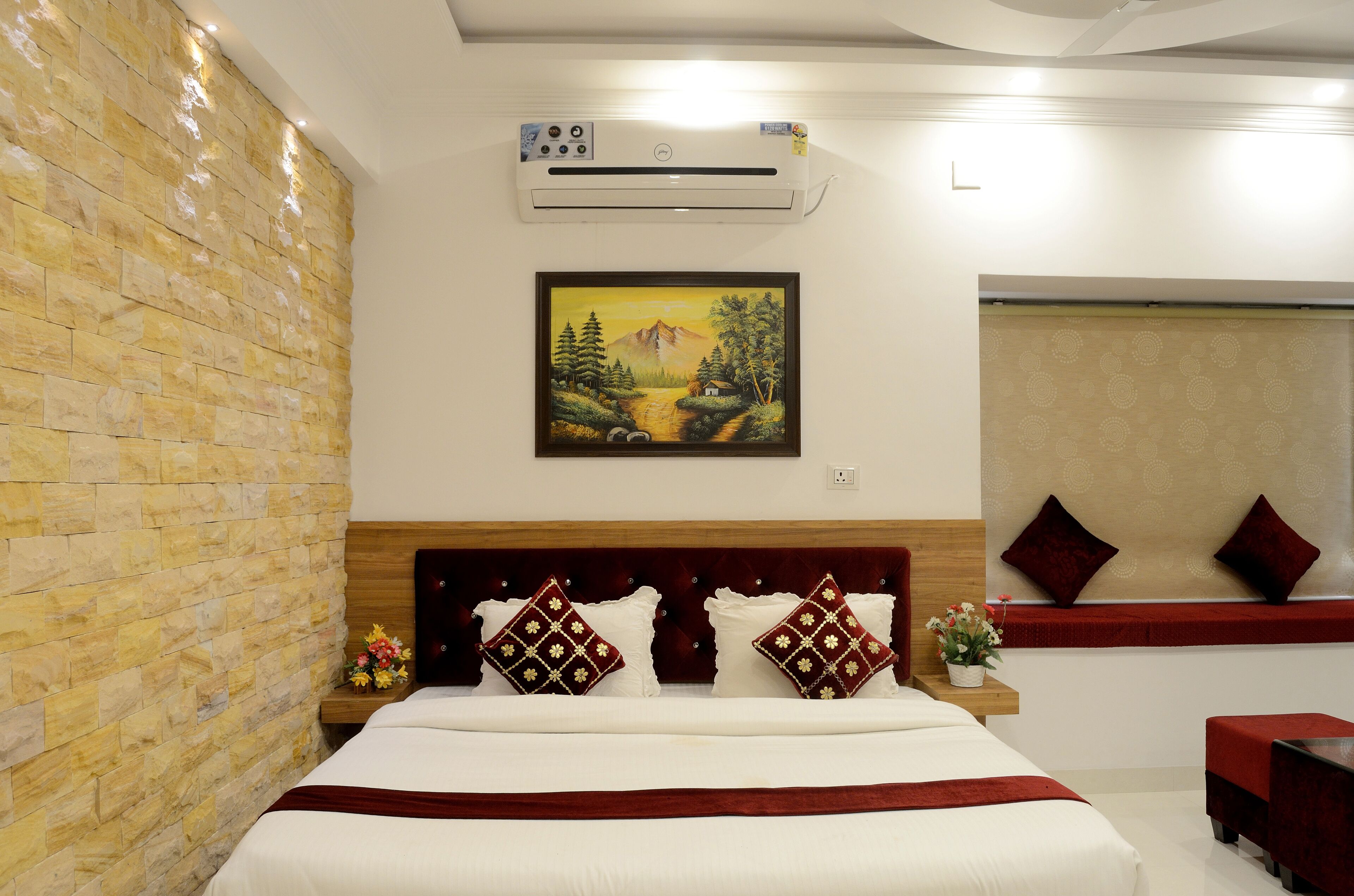 A hotel room with a stone wall, a bed with white bedding and red accents, a painting of a landscape, and a window with a patterned blind.