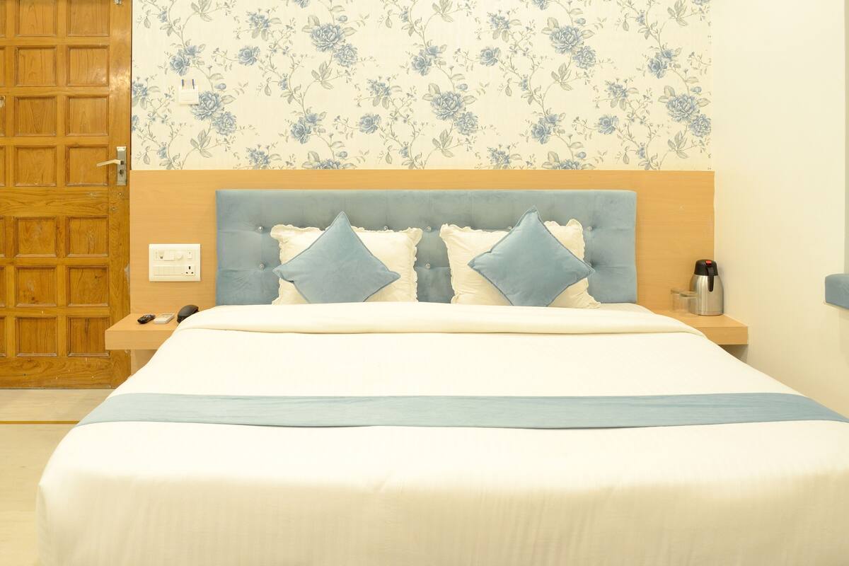 A neatly made bed with a headboard, a bedside table with a lamp, and a floral-patterned wall.