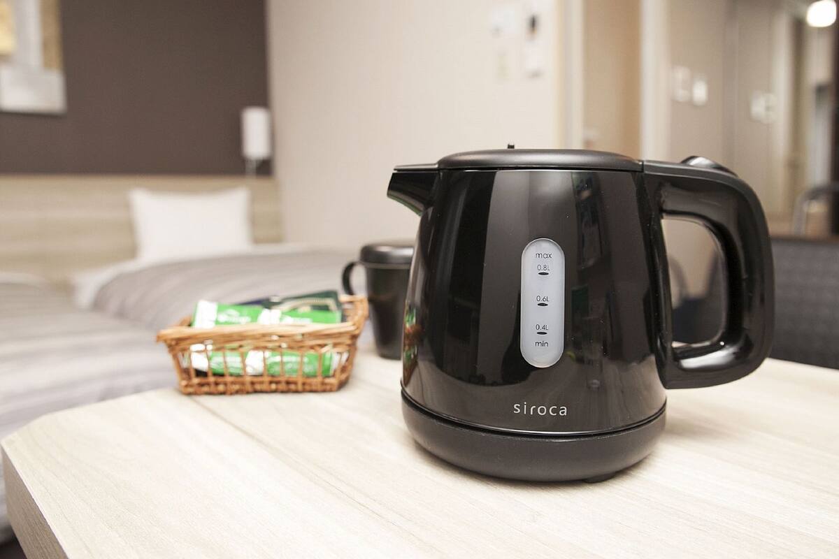 Una tetera eléctrica negra sobre una mesa de madera, con una cesta de bolsitas de té verde y una taza negra al fondo.
