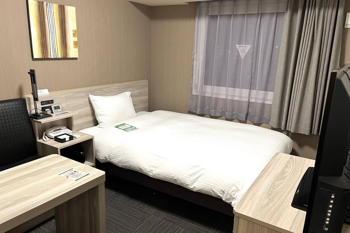 Una habitación de hotel con cama, escritorio, silla, televisión y ventana con cortinas.