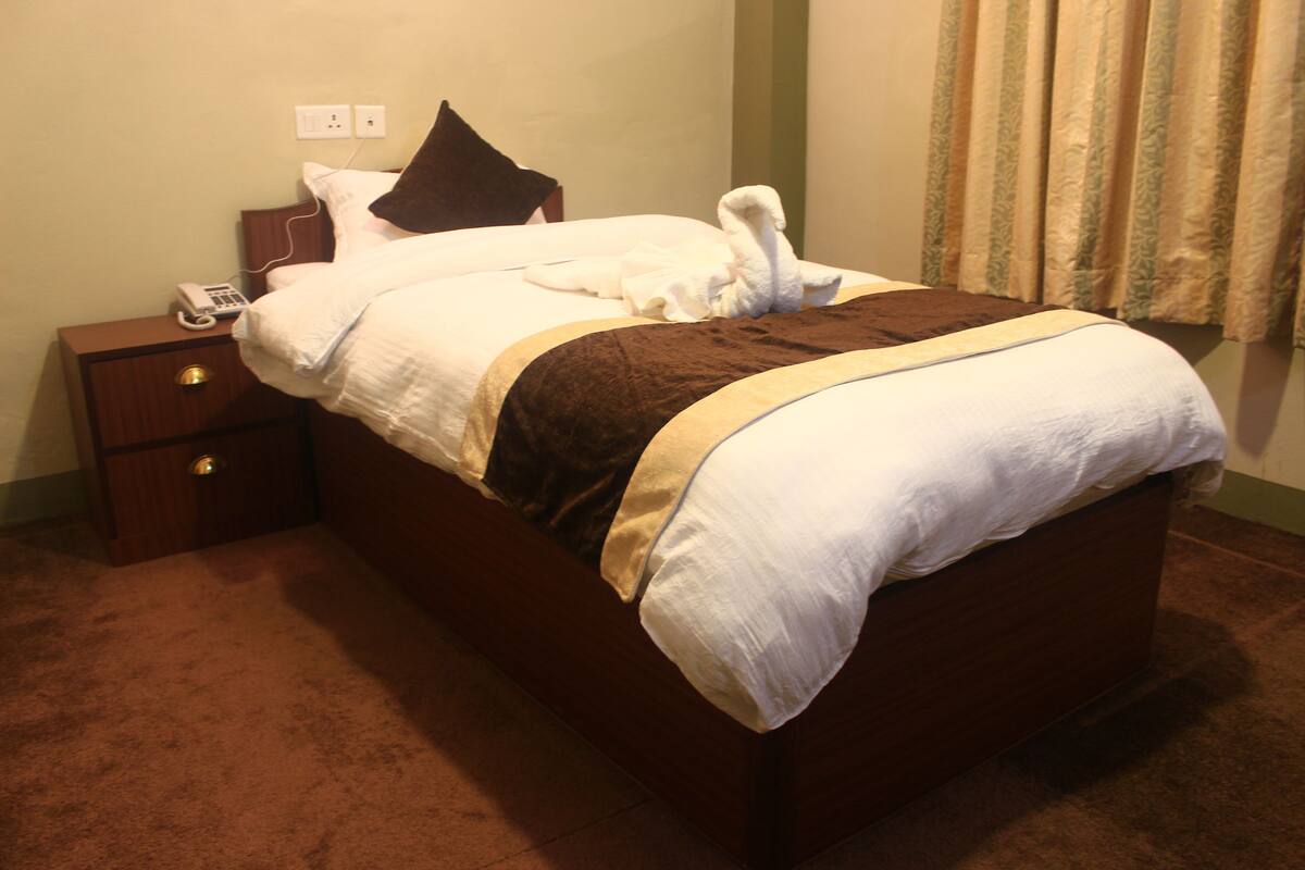 A neatly made bed with a brown headboard, a bedside table with a phone, and a power outlet on the wall.