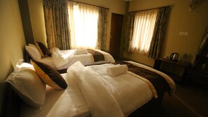 Une chambre d’hôtel avec deux lits, chacun agrémenté de draps blancs et d’éléments décoratifs marron. On y trouve une petite table avec une bouilloire et une bouteille d’eau. La chambre est équipée de rideaux et d’un miroir mural.