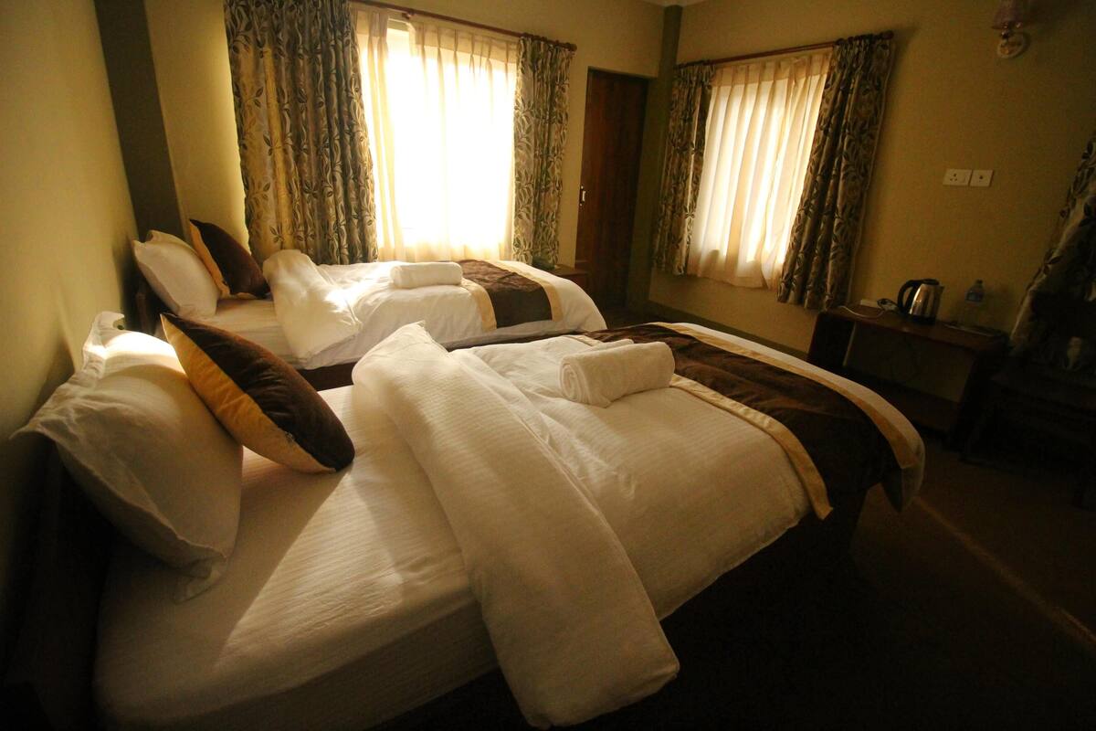 A hotel room with two beds, each with white linens and brown accents. There is a small table with a kettle and a bottle of water. The room has curtains and a mirror on the wall.
