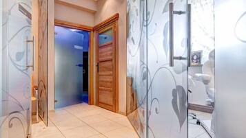 Stylish hallway with a clean design, featuring a glass door and a mirrored wall, leading to a room with a wooden door and a blue-lit interior.
