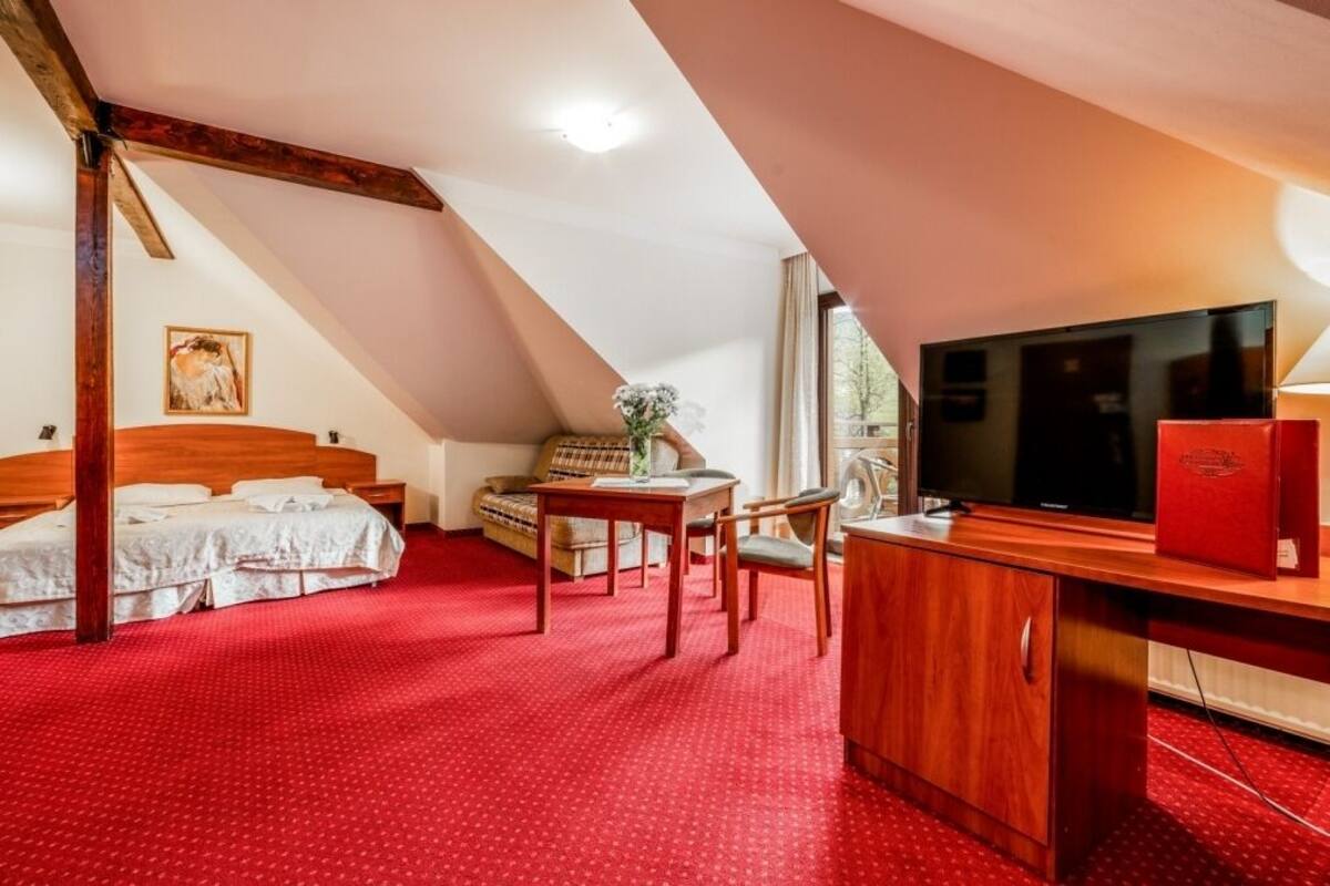 A room with a red carpet, a bed, a desk with a computer, a TV, and a window.