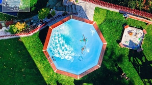 Una bañera de hidromasaje octogonal rodeada por una terraza de madera, con una piscina pequeña y un jardín con una cerca roja.
