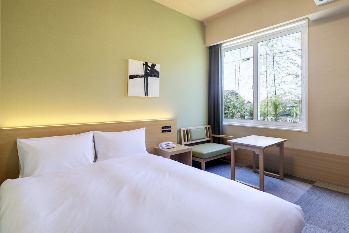 Habitación de hotel con una cama grande, una mesita de noche con un teléfono, una silla pequeña y una ventana con vistas a árboles.