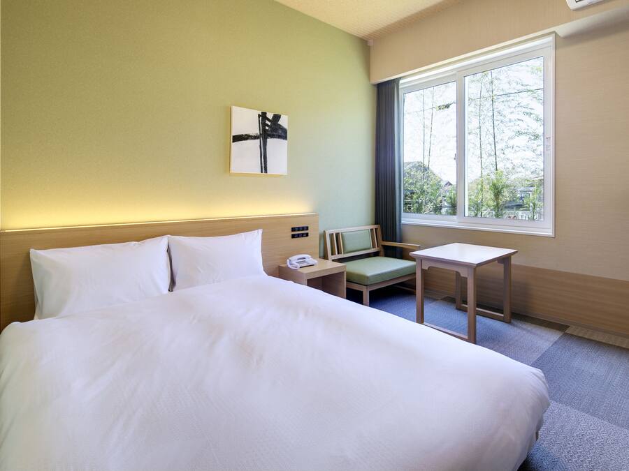 A hotel room with a large bed, a bedside table with a phone, a small chair, and a window with a view of trees.