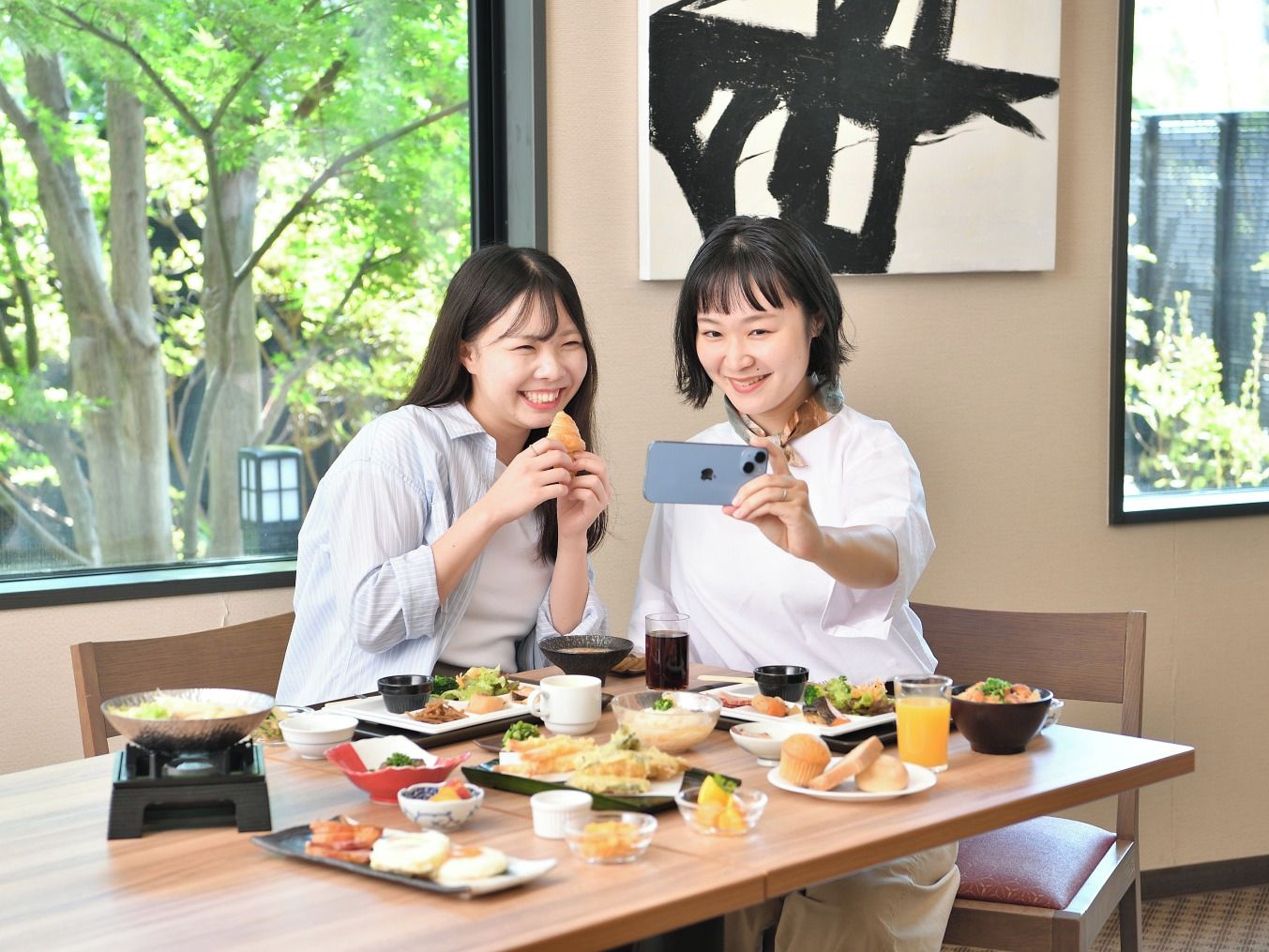 Two individuals seated at a table with various dishes, a smartphone, and a large window with a view of trees.