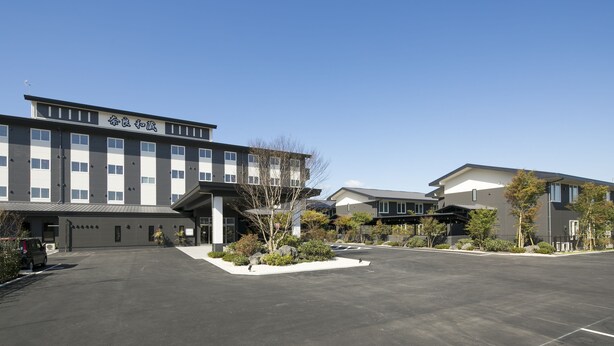 A modern building with a flat roof, large windows, and a parking area with trees and shrubs. - Grandvrio Hotel Nara Wakura (Tenri)