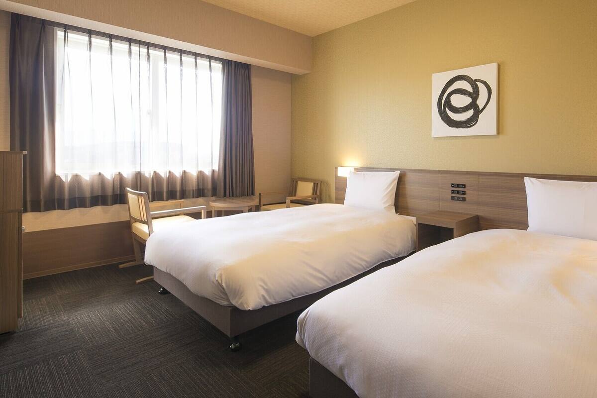Habitación de hotel con dos camas, un escritorio, una silla, un ventanal con cortinas y una obra de arte colgada en la pared.