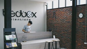 A reception desk with a modern design, a brick wall, and a clock on the wall. - Bedbox Vientiane Hostel (Vientiane)