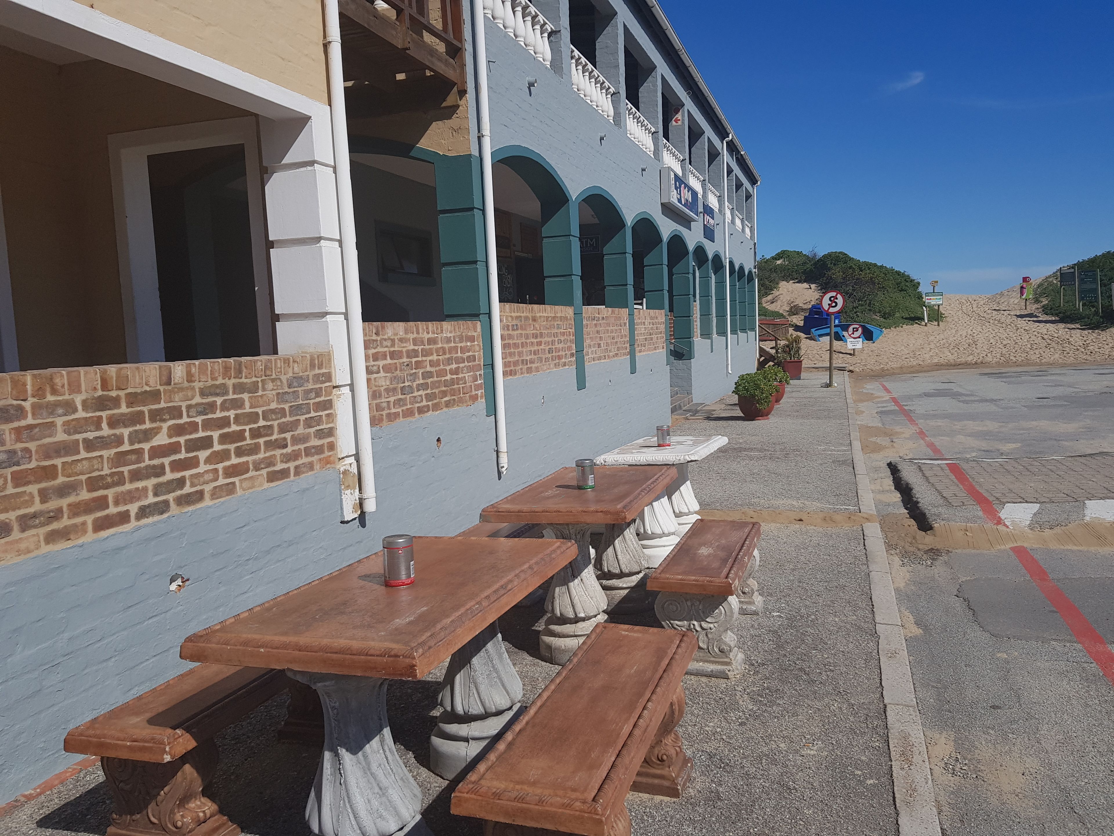 An outdoor seating area with wooden tables and benches, a brick wall, and a building with arched windows and a sign.