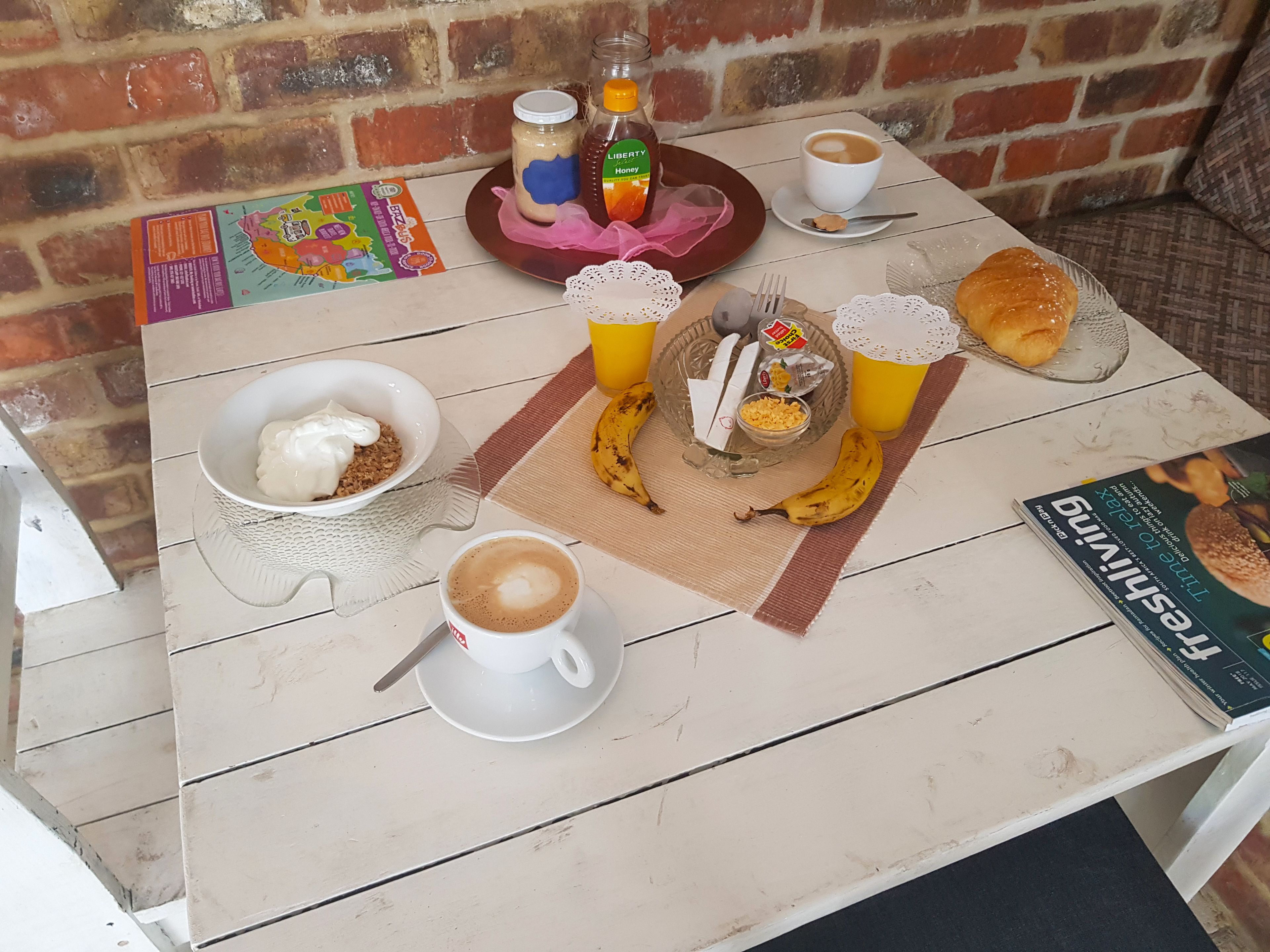 Une table dressée avec un bol de yaourt, une tasse de café, une banane, un pot de miel, une bouteille de miel, une miche de pain et un livre.