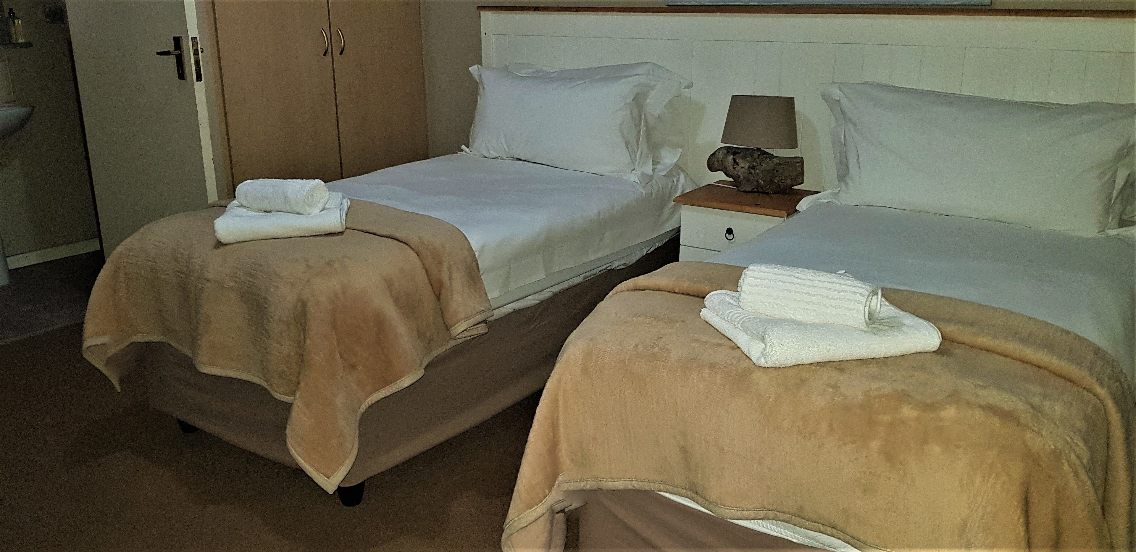 A hotel room with two beds, each with a beige blanket and white pillows, a nightstand with a lamp and a decorative item, and a wooden wardrobe.
