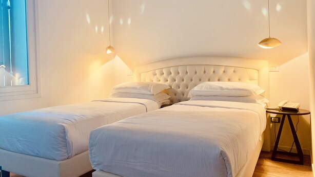 A hotel room with two beds, a bedside table with a lamp, and a window.