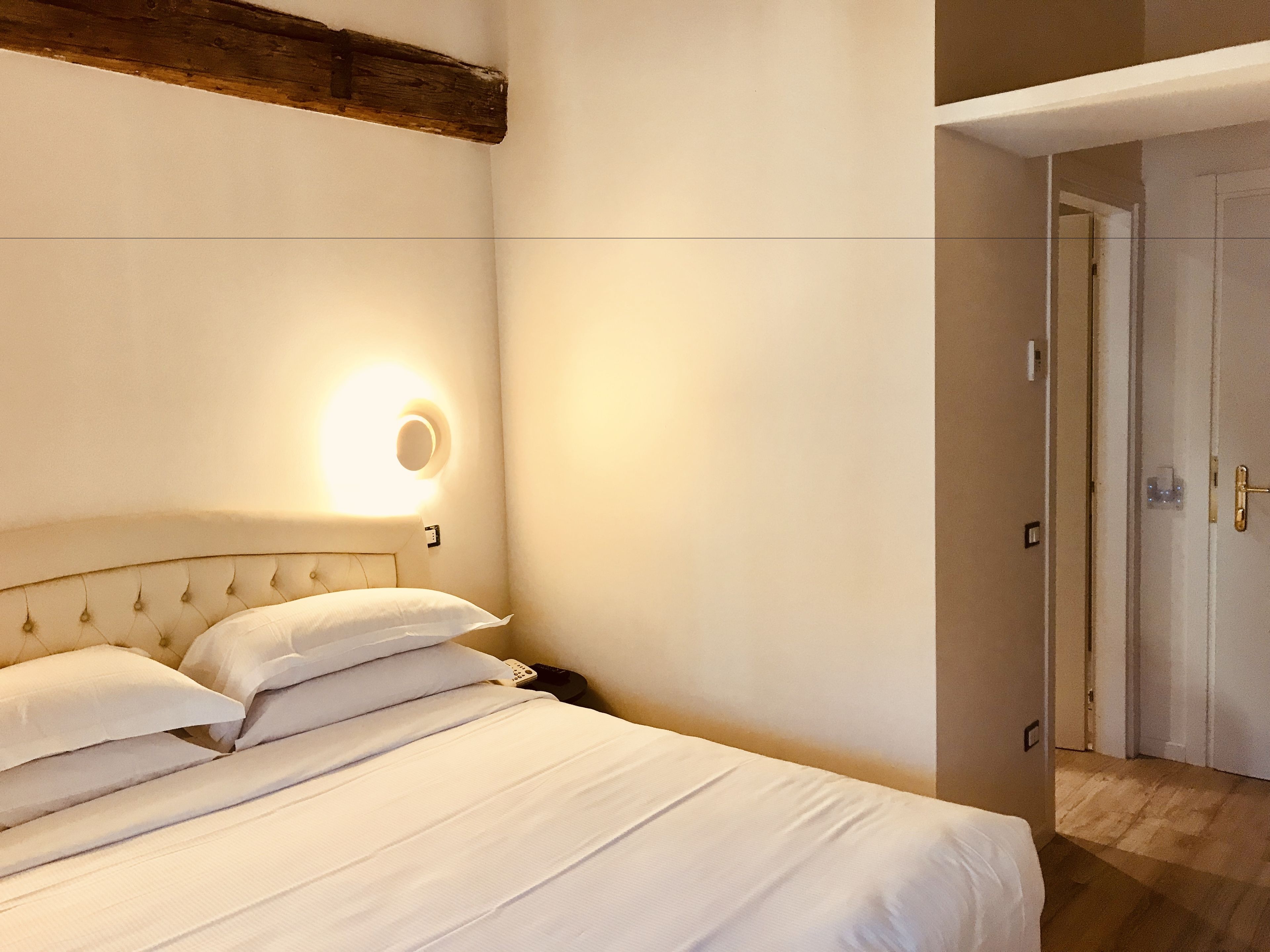 A neatly made bed with white linens, a round wall light, and a wooden headboard.