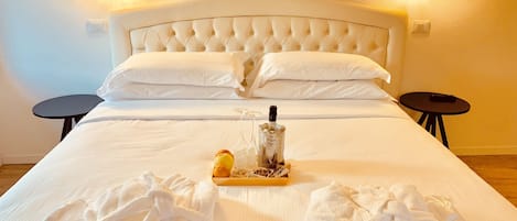 A neatly made bed with white linens, two pillows, and a tray with a bottle and glasses. There are two small round tables beside the bed.
