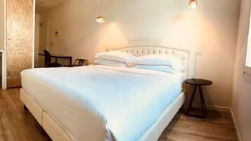 Una cama bien hecha con cabecera blanca, una mesita redonda pequeña y una silla en una habitación con suelo de madera y pared lisa.