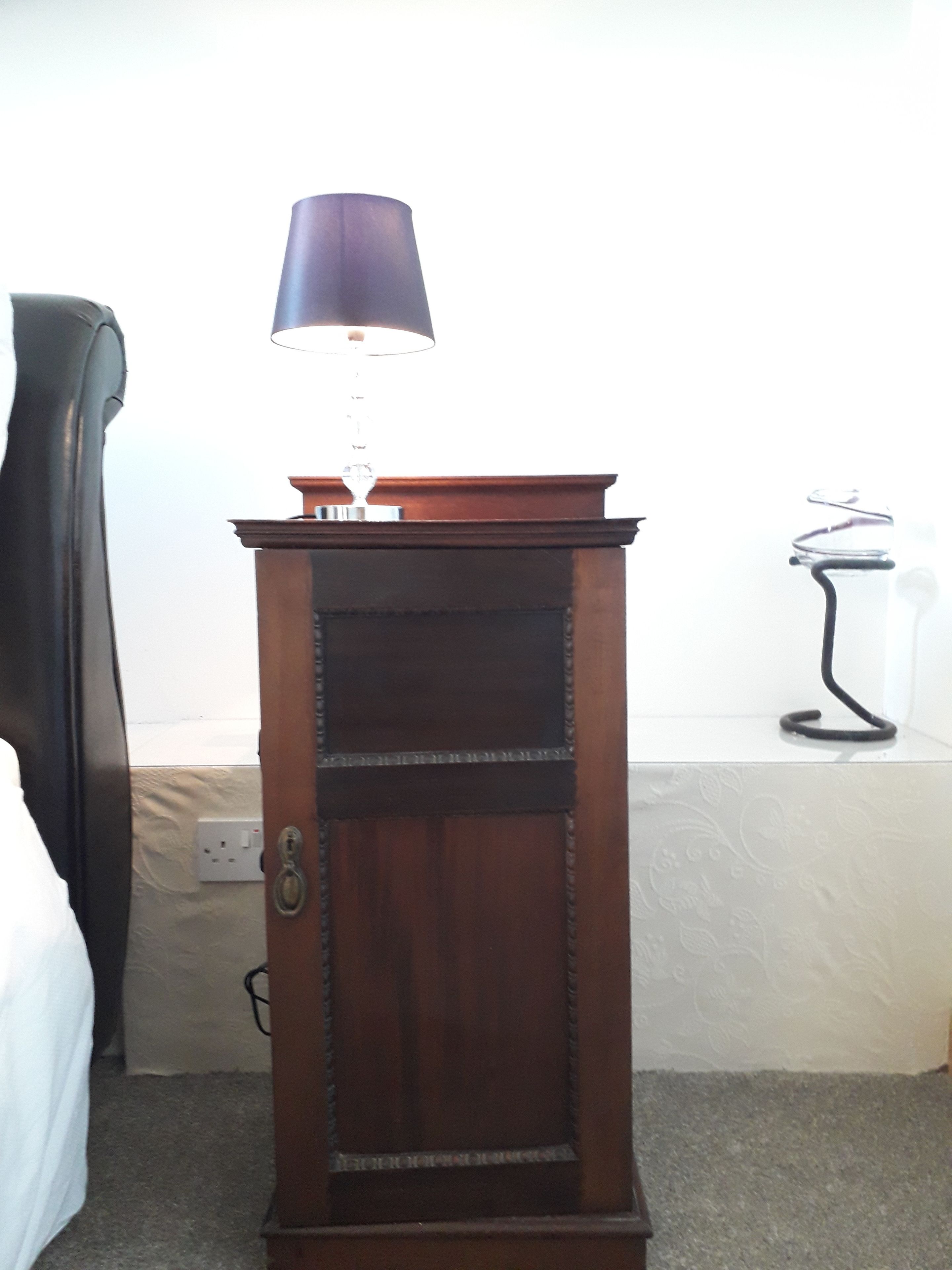 A wooden cabinet with a lamp on top, a power outlet, and a bed in the background.