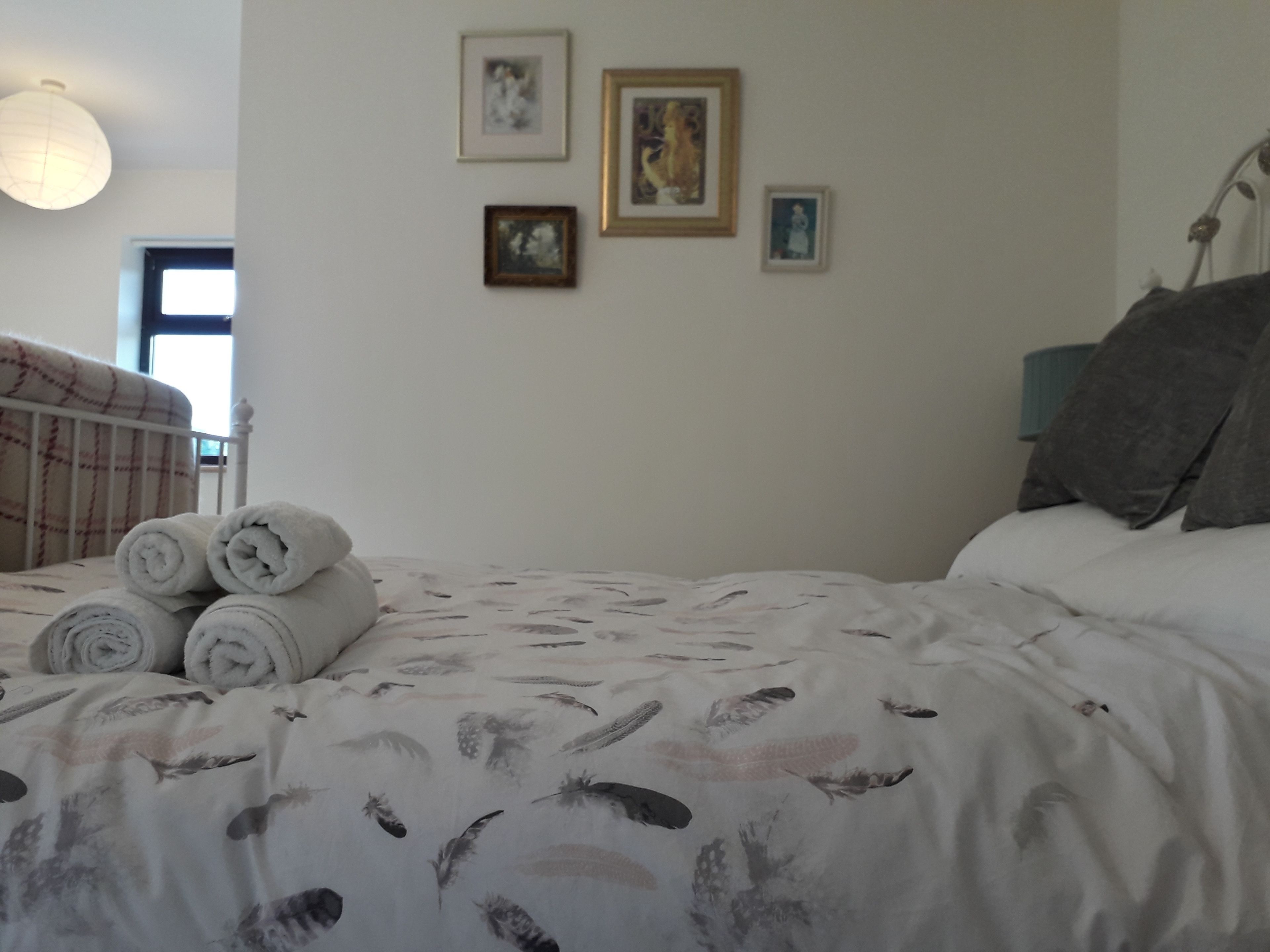 A neatly made bed with rolled towels, a plain wall with three framed pictures, and a window with blinds.