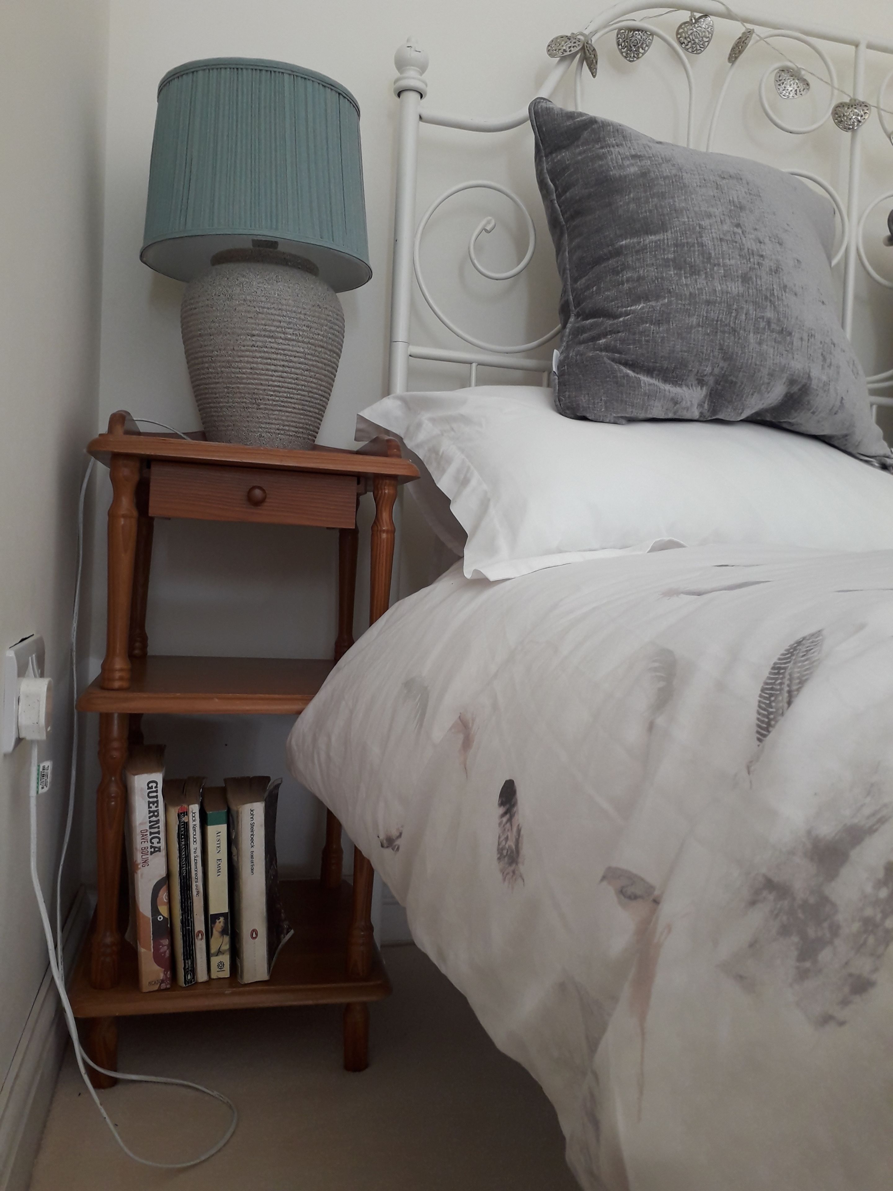 Cama bem arrumada com edredom branco e travesseiro cinza, mesa de cabeceira de madeira com luminária e livros, e cabeceira metálica branca.