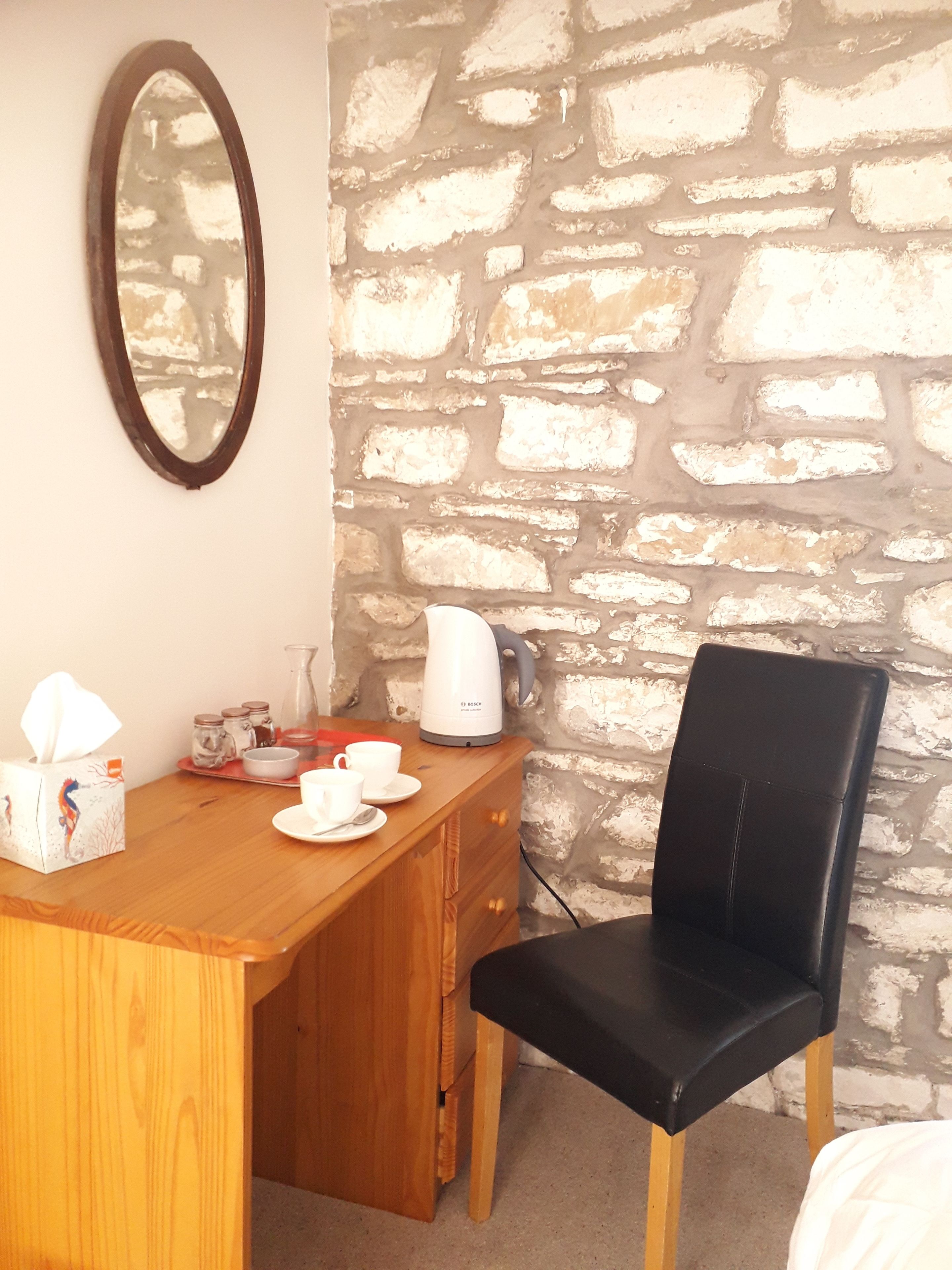 A wooden desk with a mirror, a coffee maker, and a chair in a room with a stone wall.