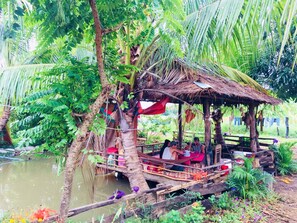 A rustic wooden structure with a thatched roof, surrounded by lush greenery and a body of water. - Family Batcave Homestay - Hostel (Banan)