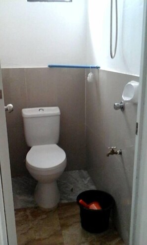 Un baño pequeño con inodoro, zona de ducha y un cubo negro en el suelo.