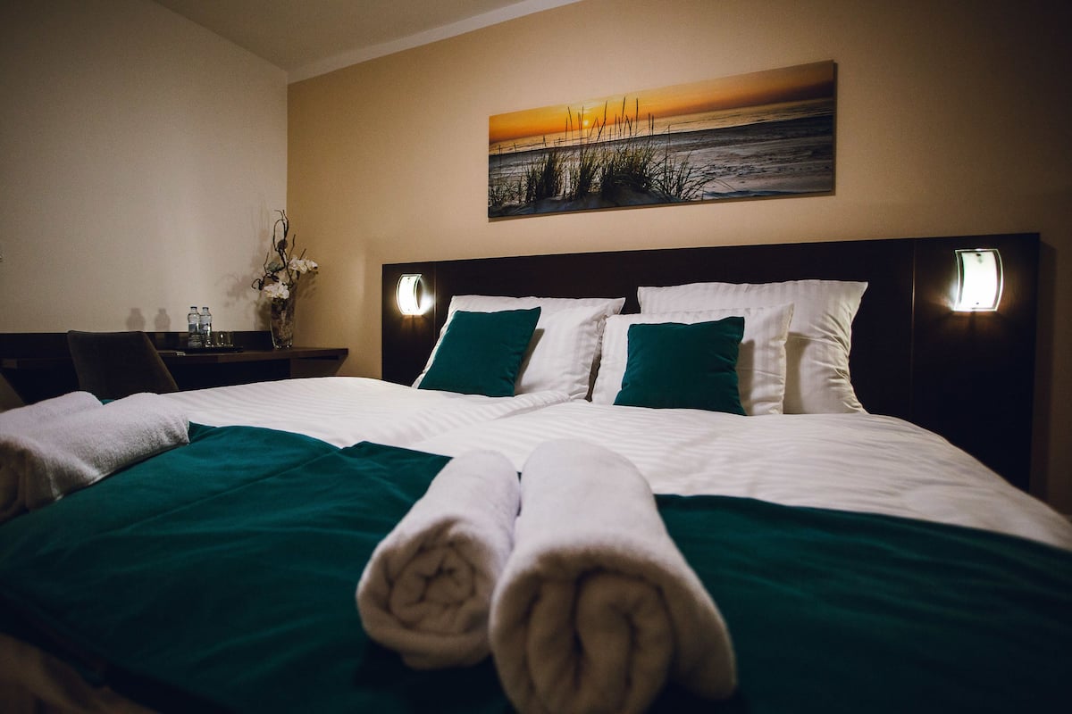 A hotel room with a neatly made bed, green and white bedding, a painting of a sunset over the ocean on the wall, and a nightstand with a vase of flowers.