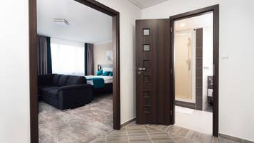 Habitación de hotel con una puerta de madera oscura, una cama con ropa de cama azul, un sofá oscuro y un baño visible a través de la puerta.