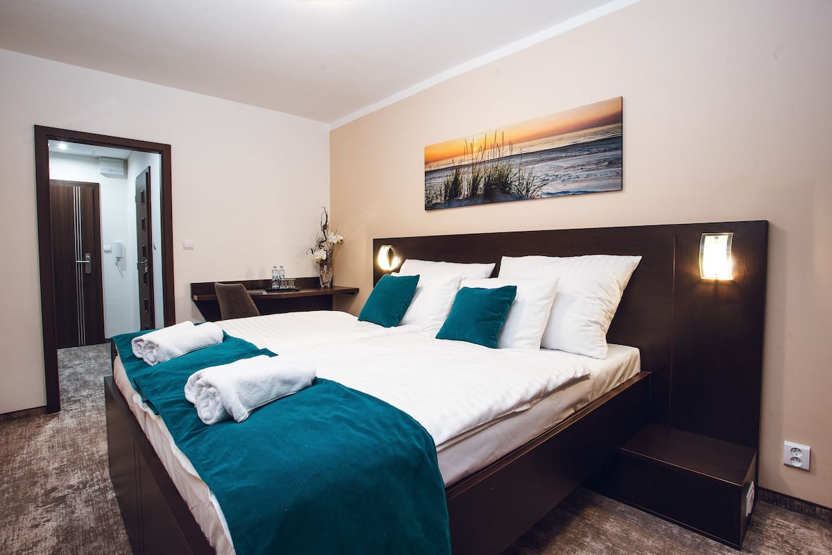 A neatly made bed with white and teal bedding, a bedside table with a lamp, and a painting of a sunset on the wall.