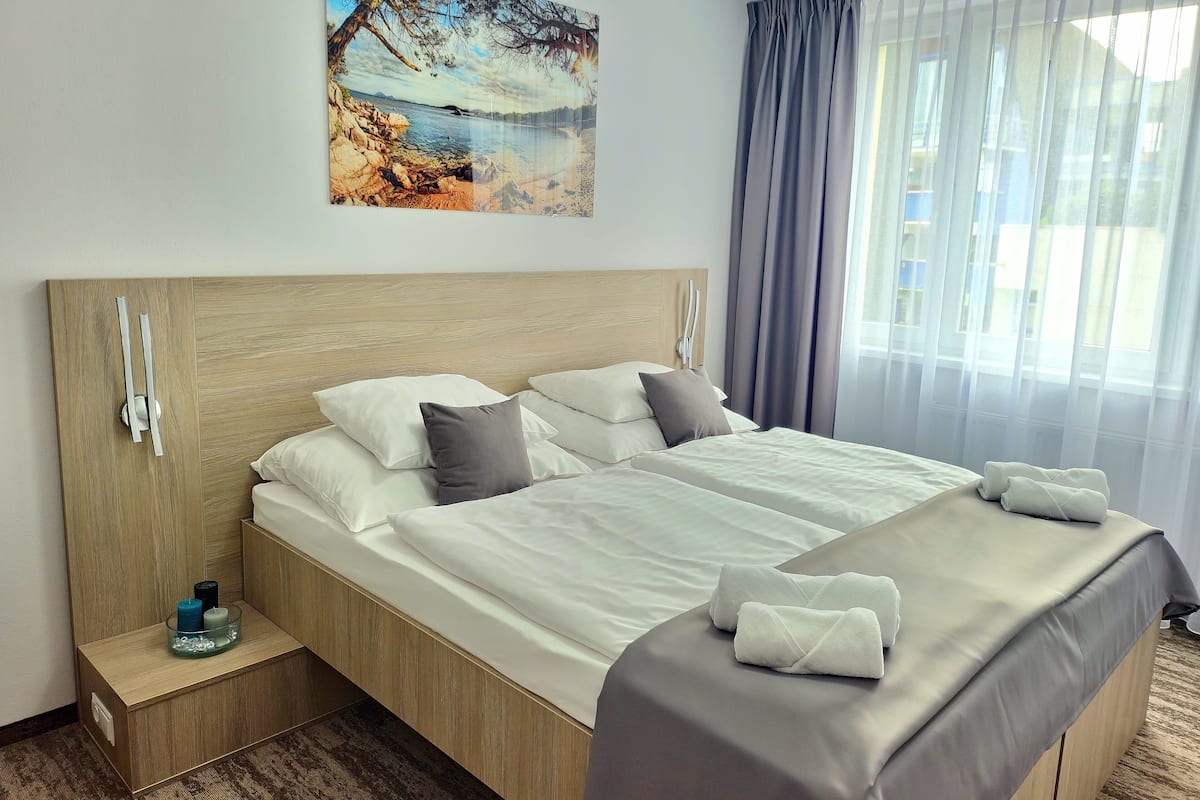 A hotel room with a large bed, a wooden headboard, a bedside table with a glass container, and a painting of a beach scene.