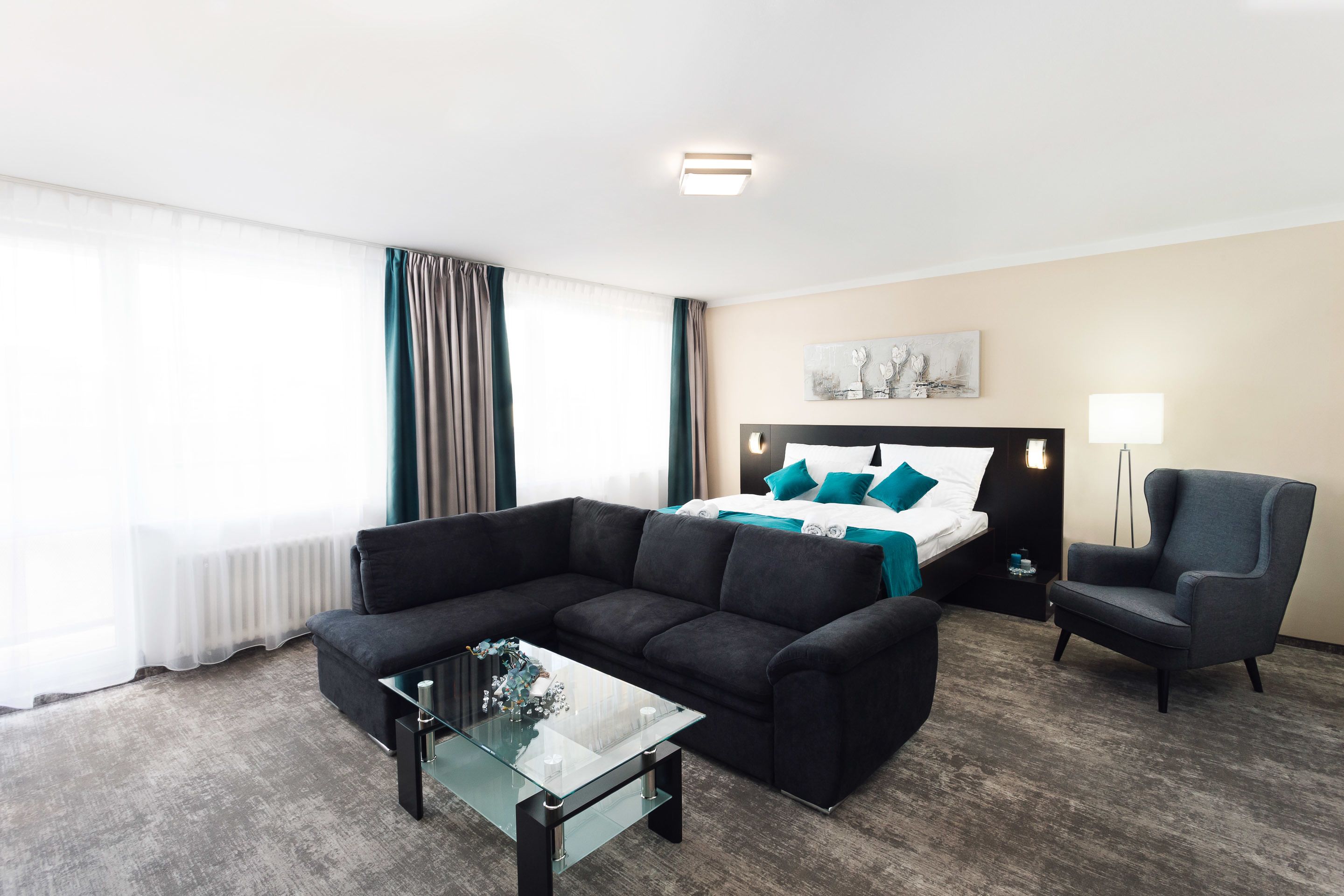 A hotel room with a bed, a sofa, a chair, a glass coffee table, and a lamp.