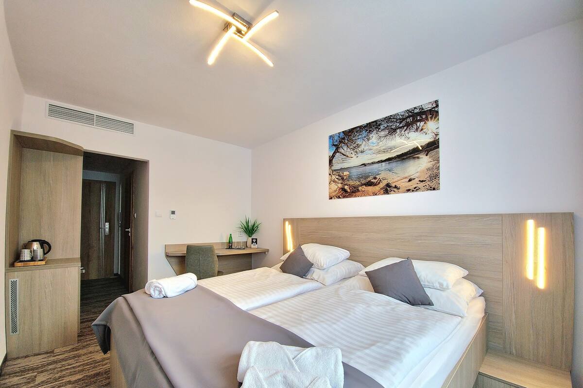 A hotel room with a large bed, a desk, a chair, a painting of a beach scene, and a door leading to another room.