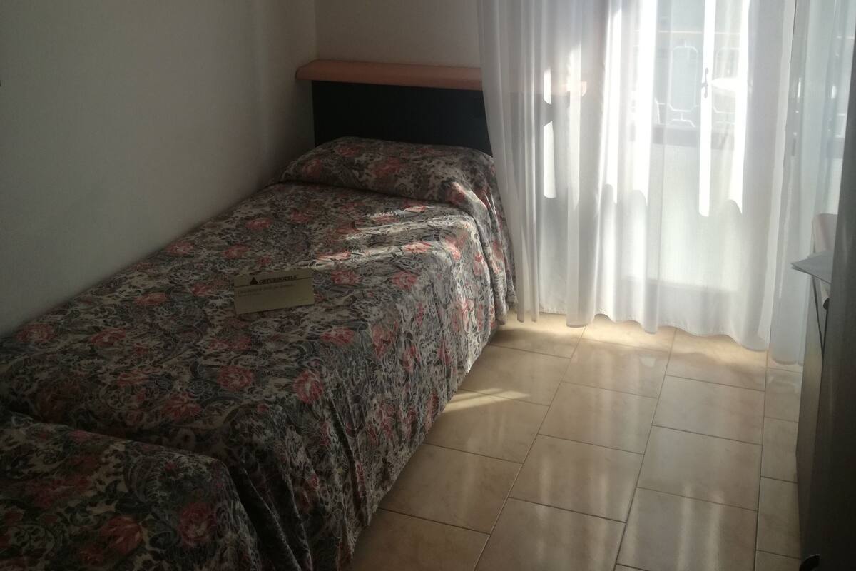 A small, single-bed room with a floral bedspread, a wooden headboard, and a large window with sheer curtains.