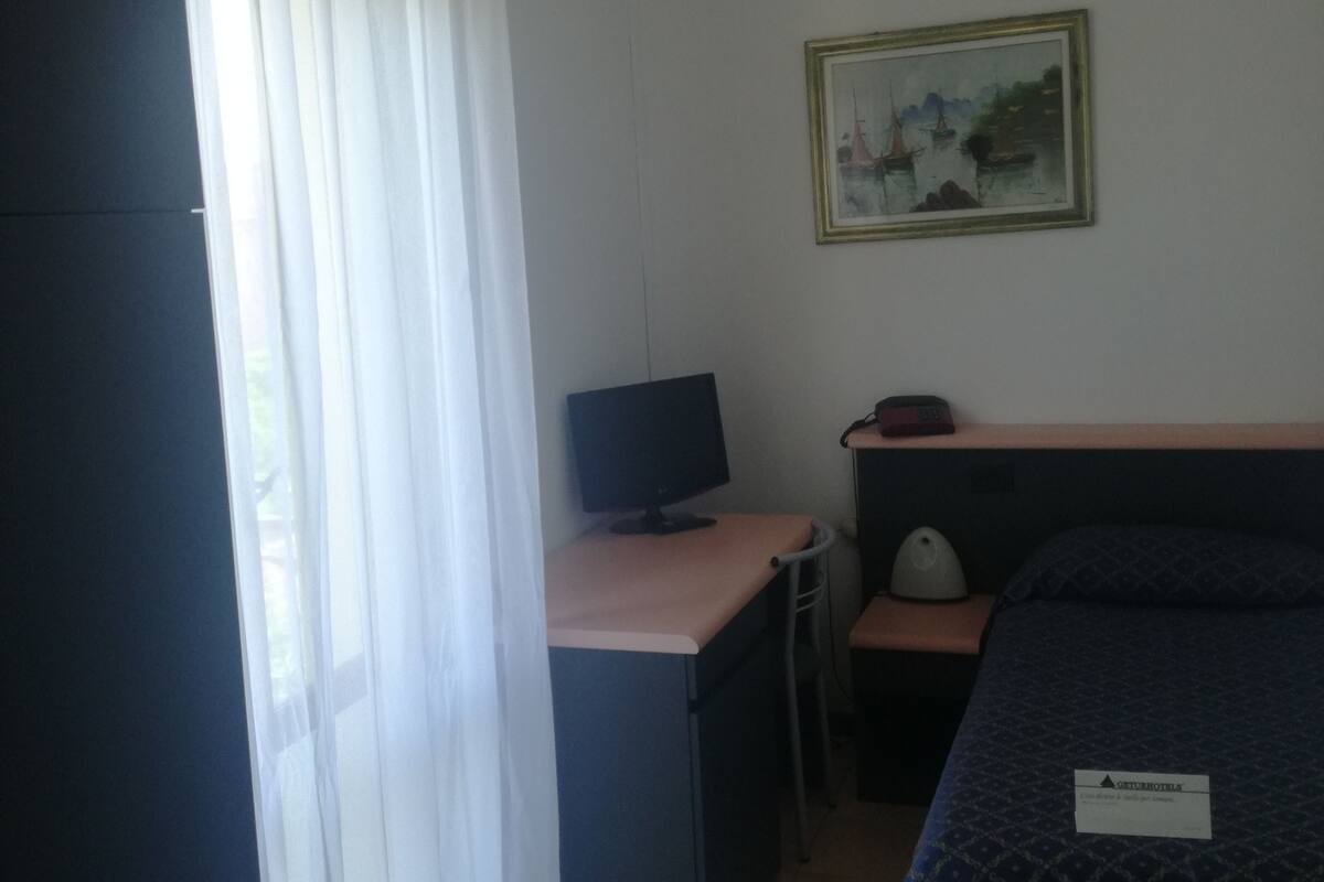 A hotel room with a bed, a desk with a computer, a chair, a bedside table, a lamp, and a painting on the wall.