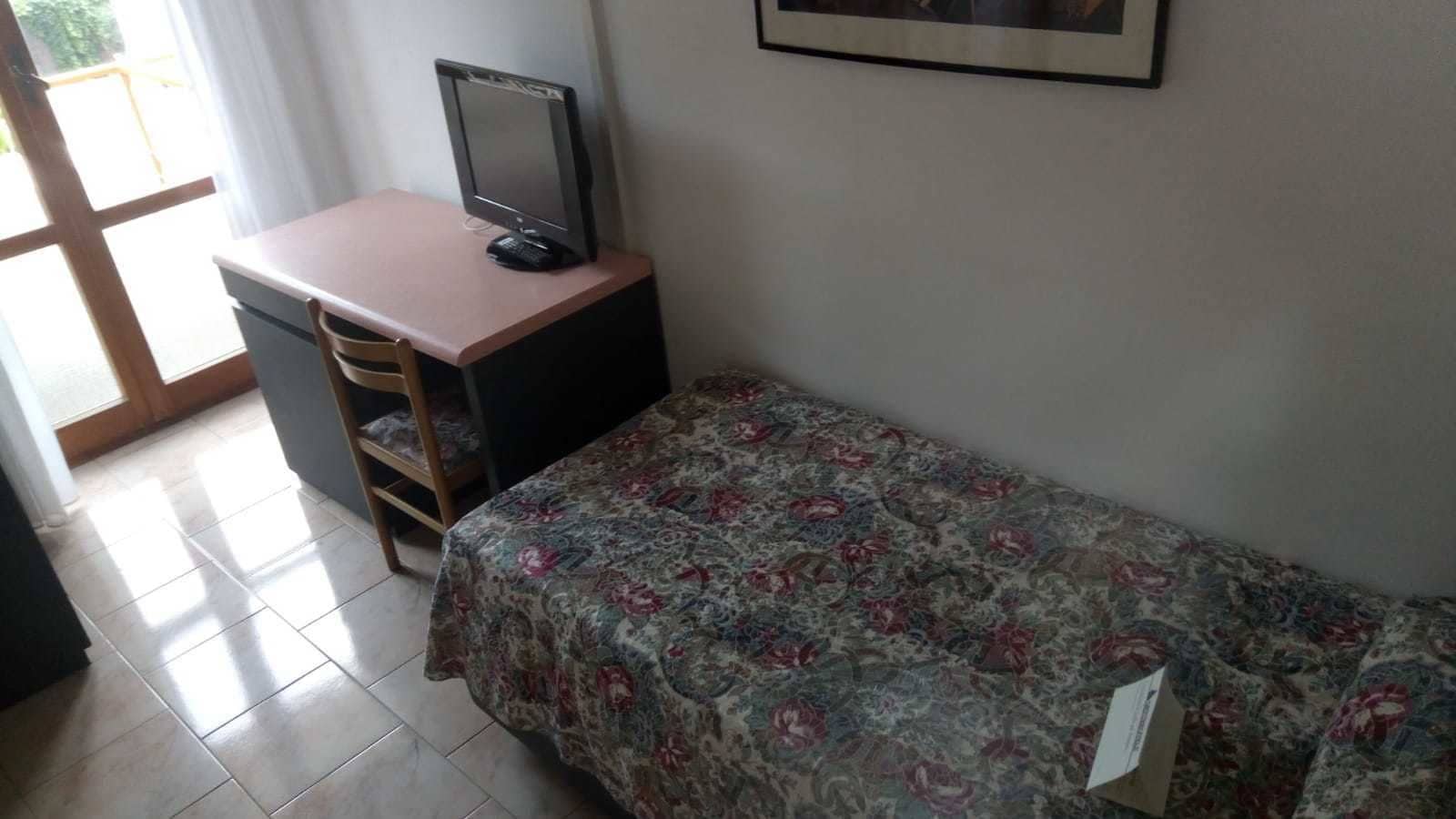 A small room with a bed, a desk, and a chair. There is a window with curtains and a door.
