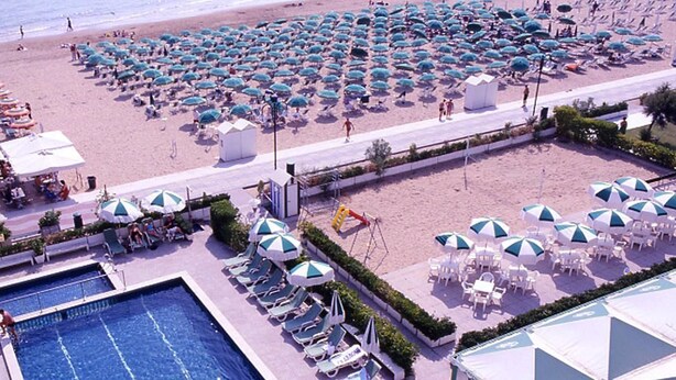An aerial view of a beach resort with a swimming pool, beach umbrellas, sun loungers, and a small building. - Hotel MaxiHeron (Jesolo)