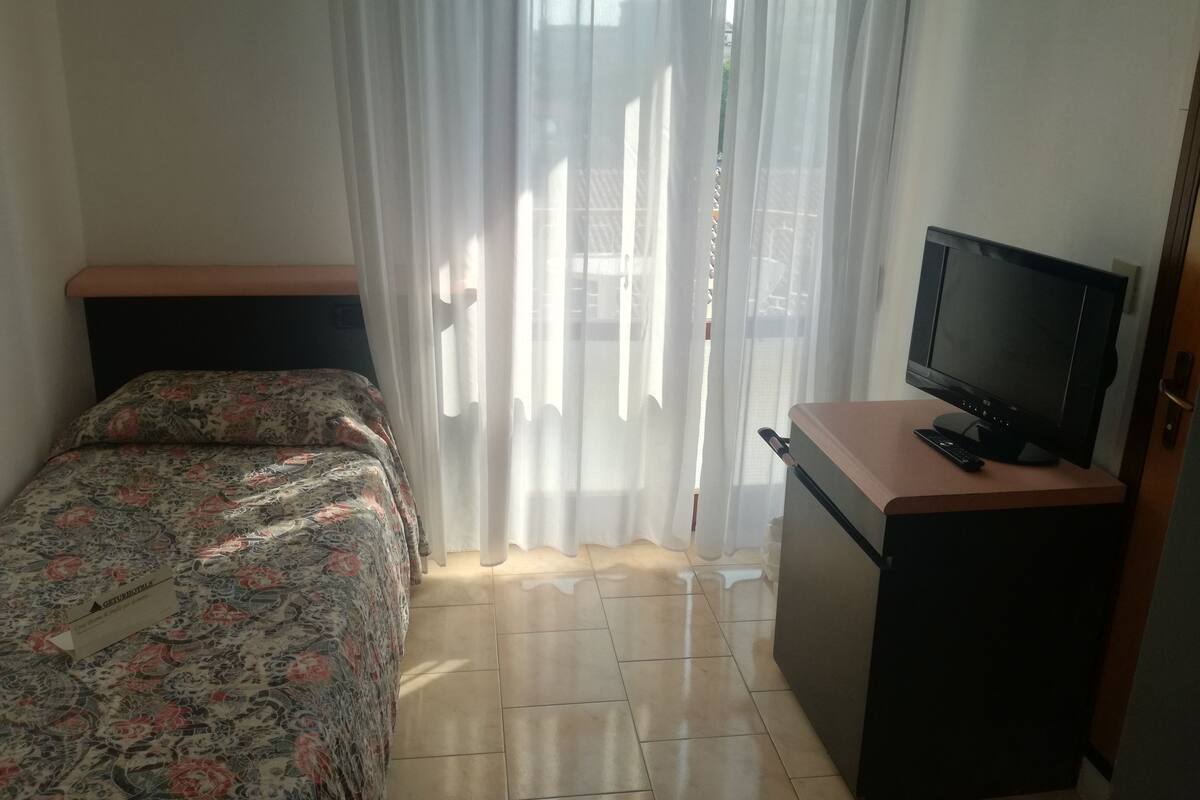 A room with a bed, a desk with a television, and a window with sheer curtains.