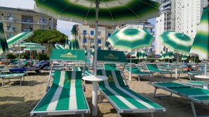 A beach with rows of green and white striped lounge chairs and matching umbrellas, some with logos, under a clear sky. - Hotel MaxiHeron (Jesolo)