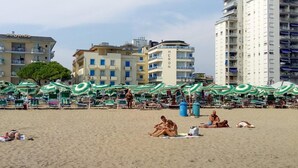 A beach with multiple green umbrellas and lounge chairs, a building labeled "FLORIDA" in the background, and a tall building with "HERON" on it. - Hotel MaxiHeron (Jesolo)