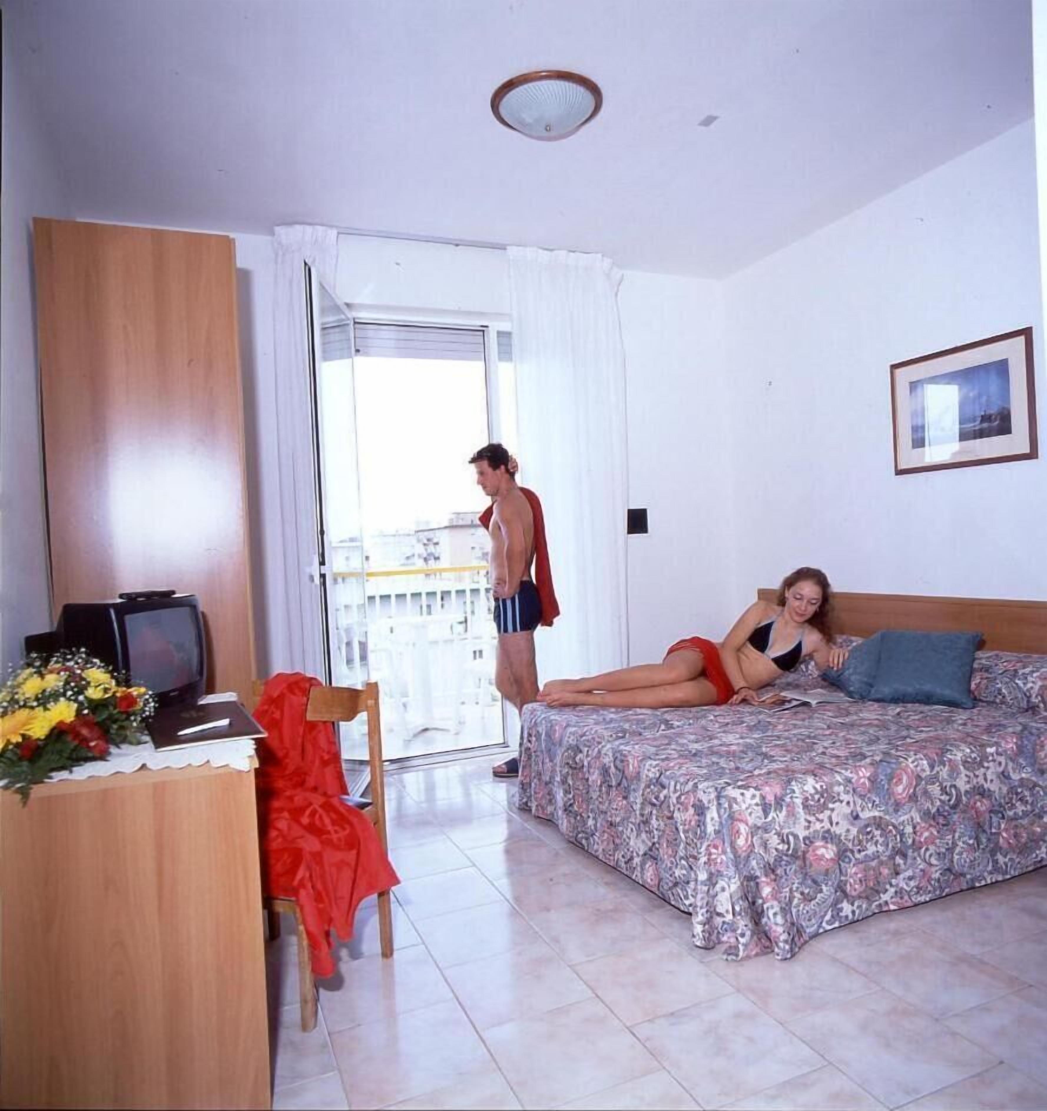 A hotel room with a bed, a desk with a TV, a chair, and a window with curtains.