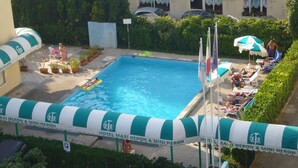 Área de piscina com espreguiçadeiras e guarda-sóis, cercada por vegetação e um edifício com varanda.